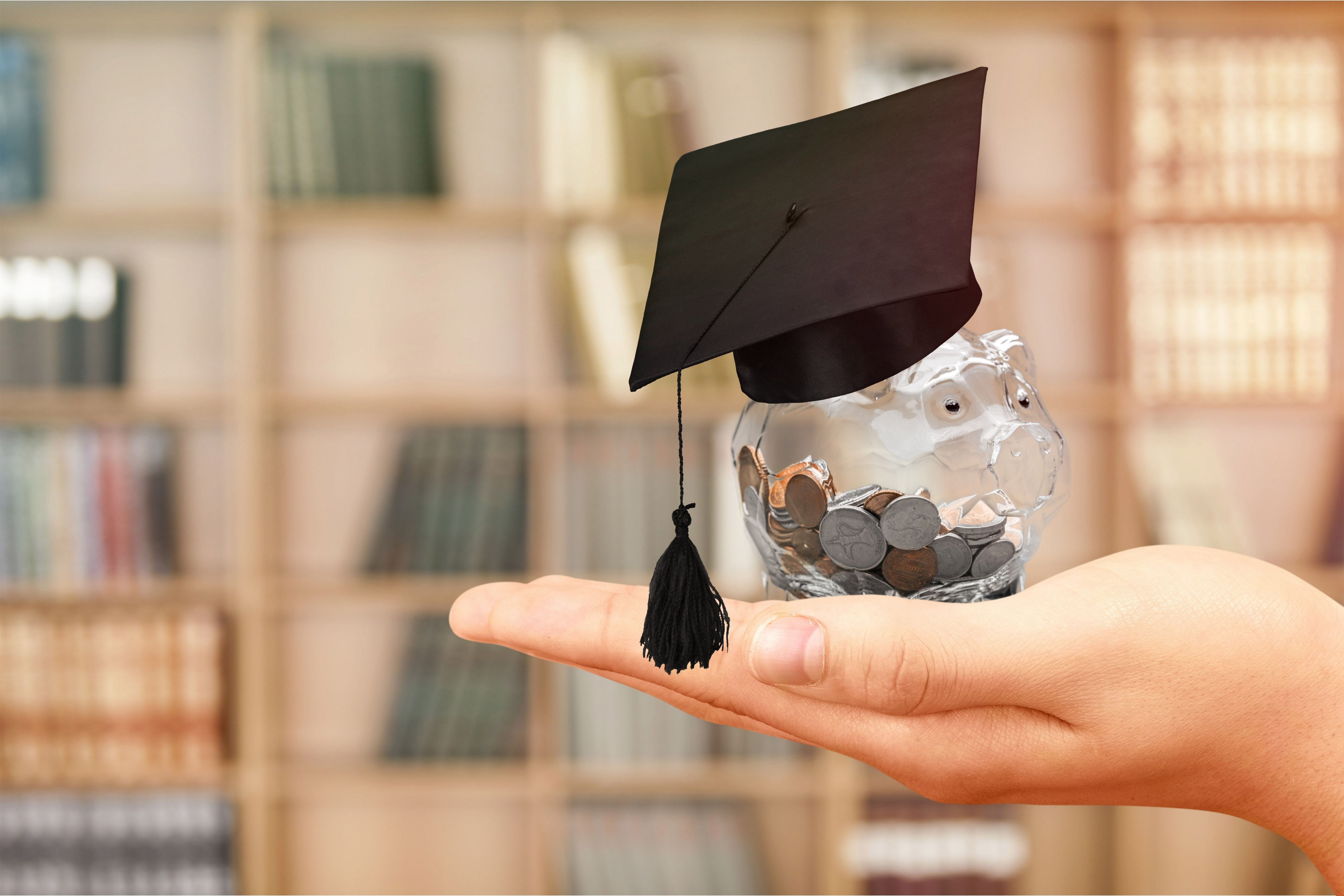 Scholarship A transparent piggy bank full of coins and with a doctoral cap is held in front of a book shelf.